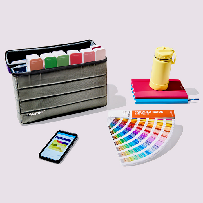 The PANTONE Formula Guide is fanned out on a desk in a colourful scene next to a mobile phone showing the PANTONE® Connect App which allows users to access over 15,000 colours in every PANTONE library. Behind the PANTONE colour guide are some notebooks and a bottle with a stylish bag containing other colour guides from the same range, including the Formula Guide, Color Bridge Set and CMYK guides.