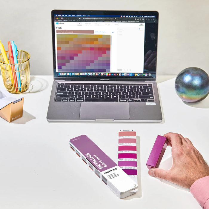 A designer comparing colours against product packaging using the PANTONE Metallics Solid Coated Guide at their desk.