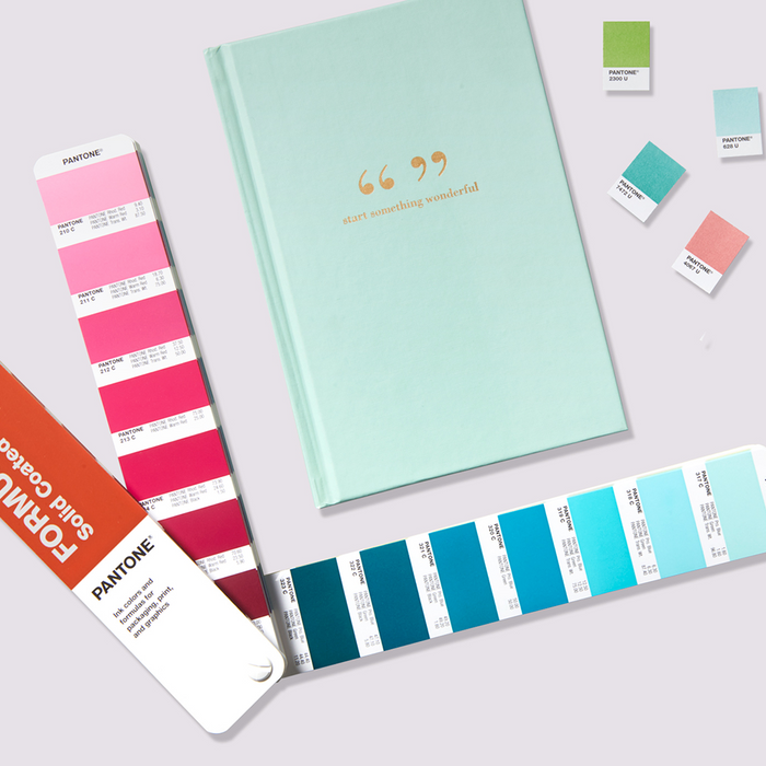 Fanned out PANTONE Formula Guide fan on a designer's desk, showing colour patches and ink mixing formulations beside colour chips and a pastel coloured inspiring notepad design.
